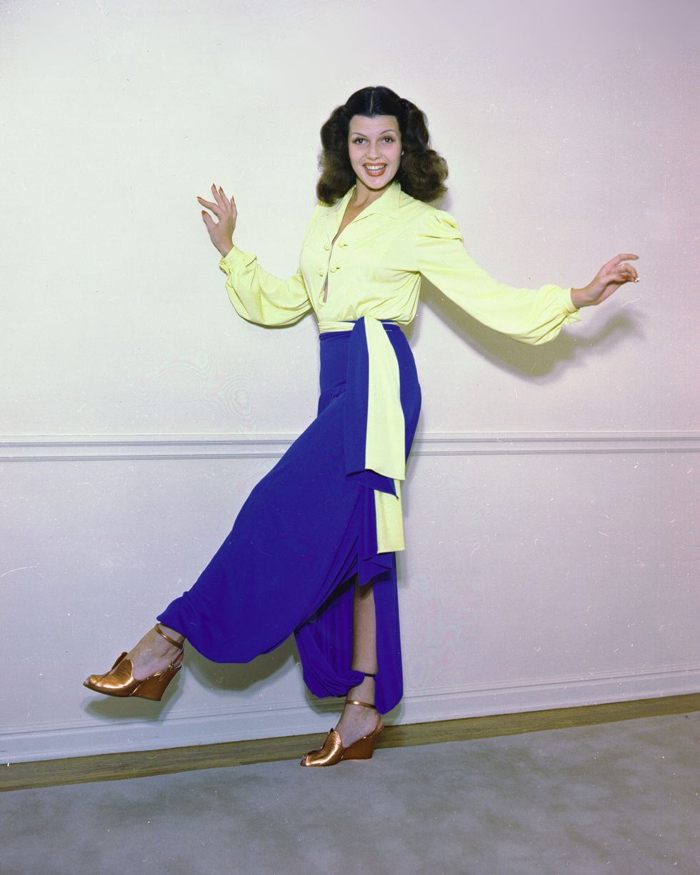 rita hayworth poses for a photo with one leg kicked slightly in the air, she holds her arms out and smiles, she wears a yellow long sleeve blouse, bright blue pants, and copper heels