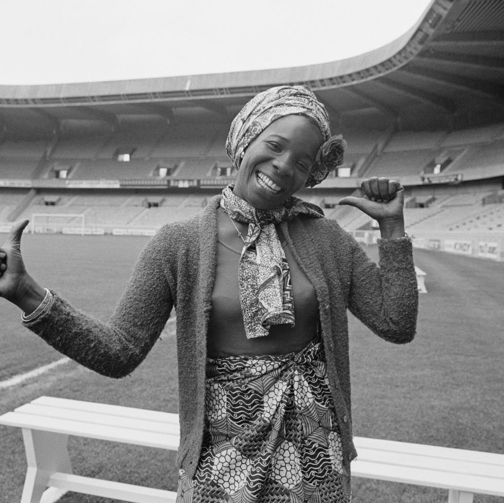 rita marley smiles at the camera and gives two thumbs up with her arms extending by her sides, she stands inside an empty stadium wearing a patterned head scarf, neck scarf and skirt with a long sleeve sweater over her shirt