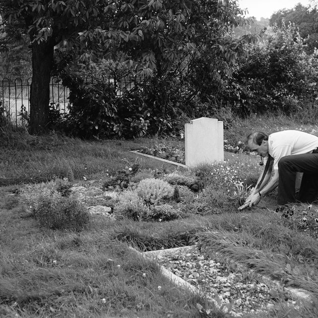 Roald Dahl smokes a pipe as he uses a pair of garden shears to tend to a grave