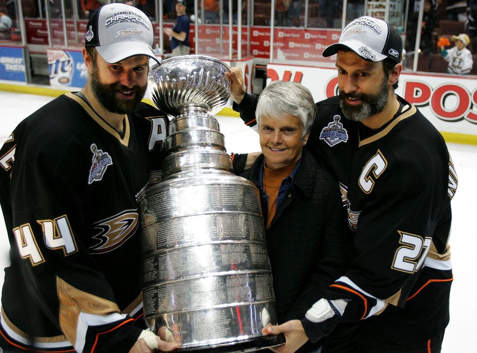 rob and scott niedermayer wear anaheim ducks hockey jersey and gray championship hats as they pose with the stanley cup, with their mother carol standing between them next to the trophy