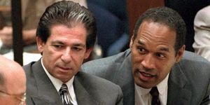 robert kardashian and oj simpson sit at a table and look at a man in the lower left corner of the frame, both kardashian and simpson wear gray suits with light colored shirts and ties