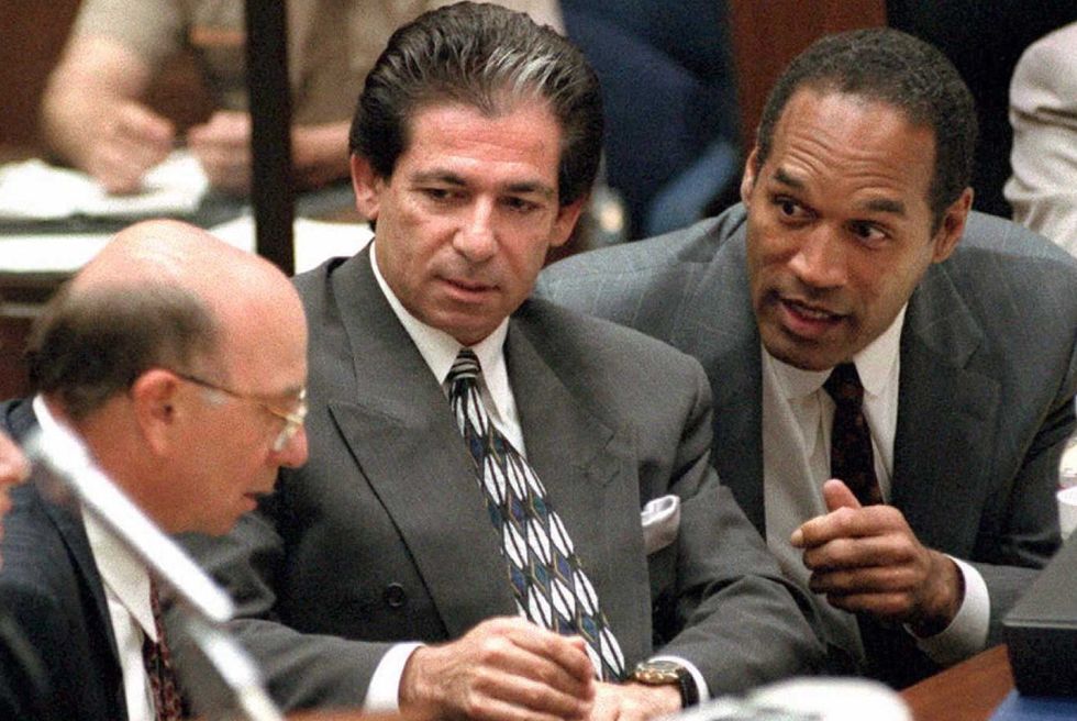 robert kardashian and oj simpson sit at a table and look at a man in the lower left corner of the frame, both kardashian and simpson wear gray suits with light colored shirts and ties