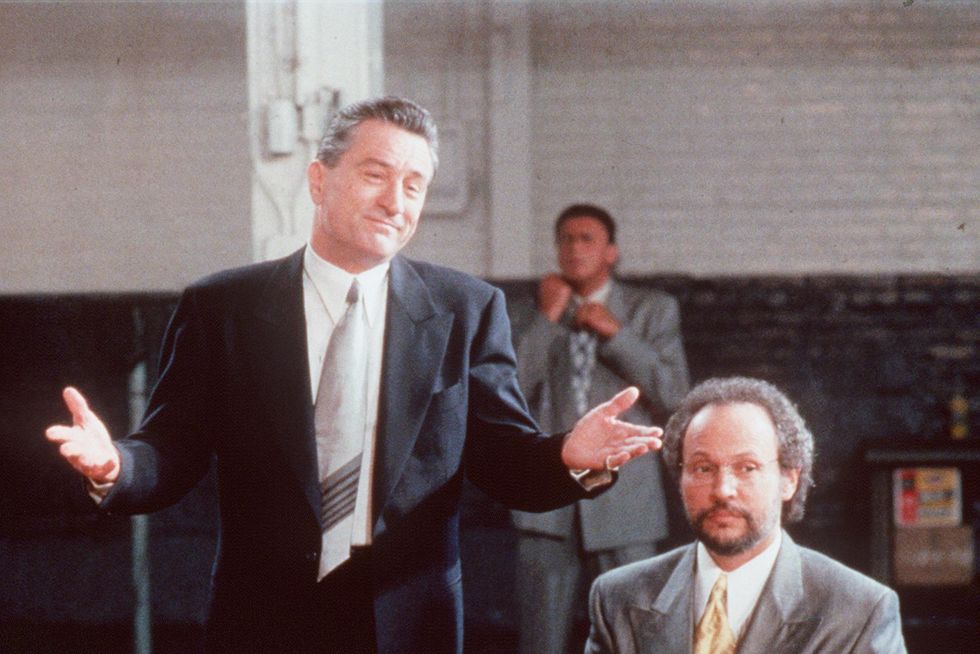 robert de niro wearing a black suit and tie and shrugging his shoulders, with billy crystal wearing a gray suit and tie and sitting next to him at a table