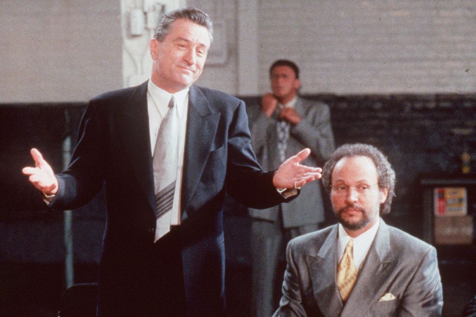 robert de niro wearing a black suit and tie and shrugging his shoulders, with billy crystal wearing a gray suit and tie and sitting next to him at a table