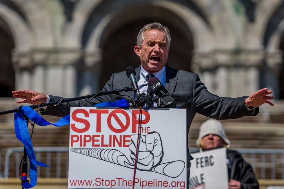 robert f kennedy jr speaks into microphones at an environmental rally, he is mid sentence and his arms are outstretched, he is wearing a gray suit with a blue collared shirt and tie, in front of him is a sign that says stop the pipeline