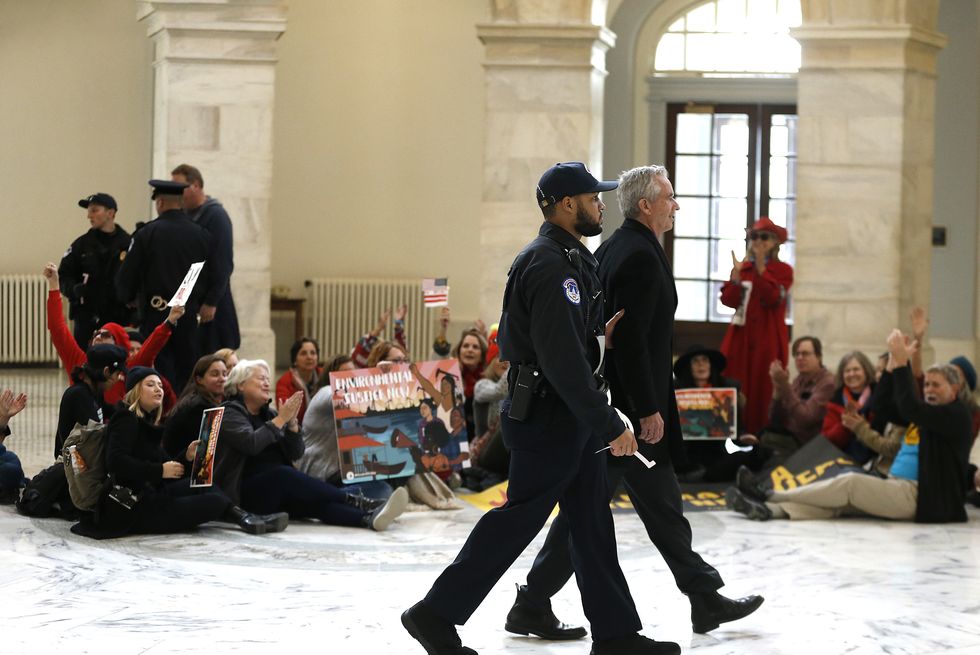 robert f kennedy jr is led by a police officer out of the us capitol building during a climate change protest, the pair is seen in profile and fellow protesters sitting on the floor in the background are applauding kennedy