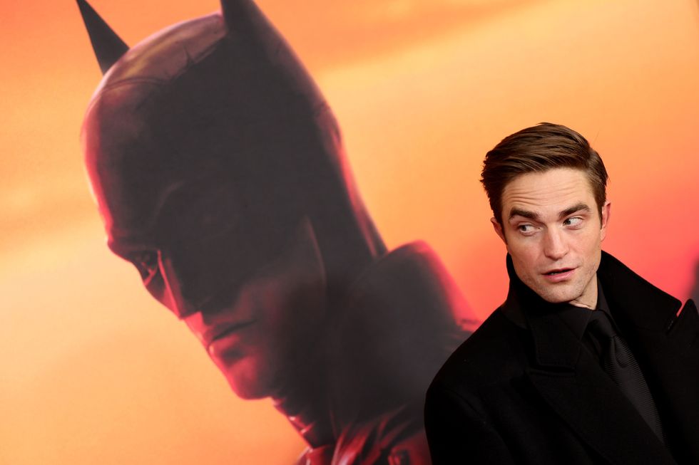 robert pattinson looks to the left as he stands in front of a large poster with a picture of himself as batman