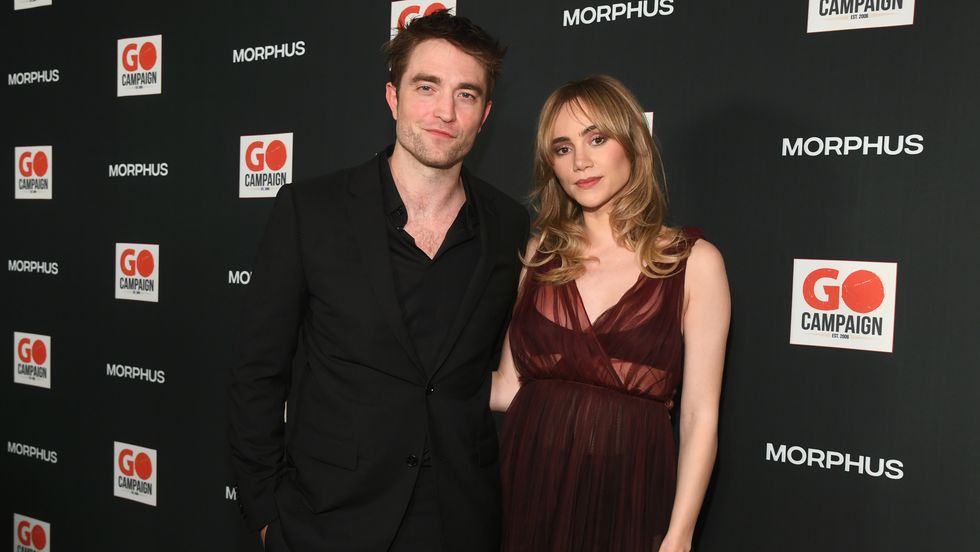 robert pattinson and suki waterhouse pose on the carpet, he's wearing a black suit and has an arm around her waist, she's wearing a see through burgundy dress