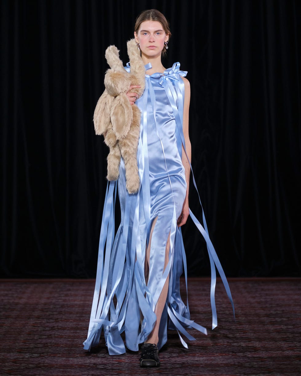 Model showcasing a blue satin dress with long ribbons and a stuffed animal