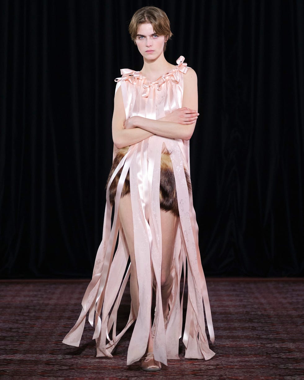 a fashion model on a runway wearing a unique outfit with flowing ribbons and fur elements