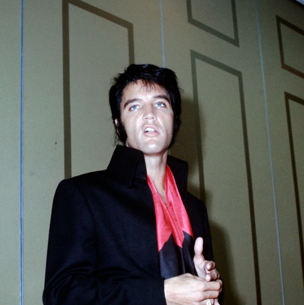 a man in a black jacket with a pink and blue shirt stands in front of a beige wall and looks to the left