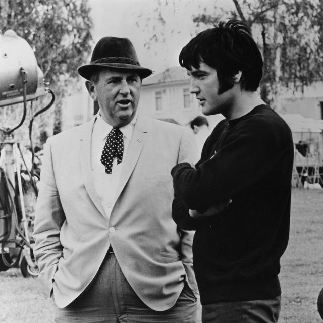 man in a suit and hat talking to another man as they stand outside with film equipment in the background
