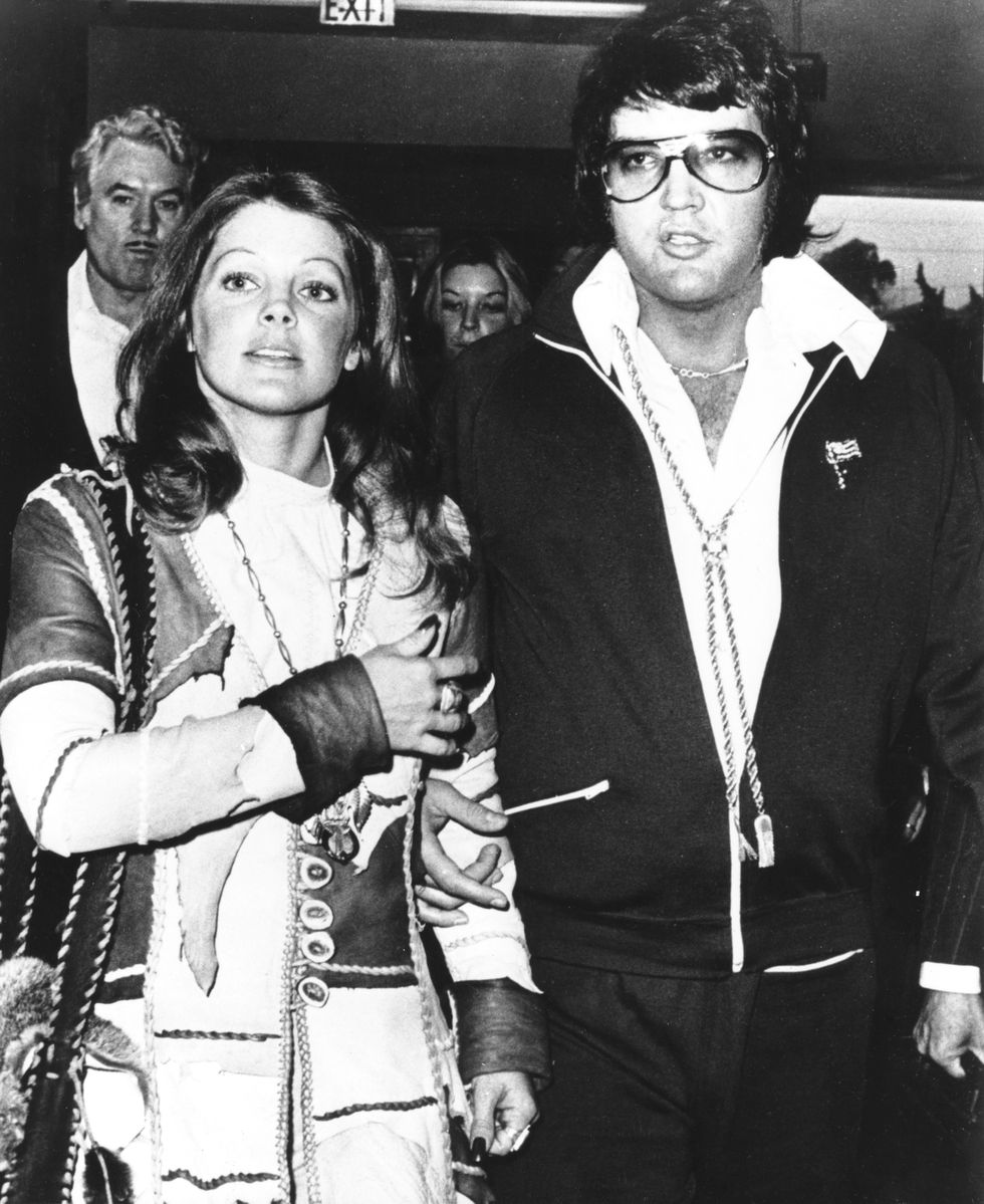 elvis and priscilla presley with their arms interlocked as they walk out of a courthouse amid a crowd of peple