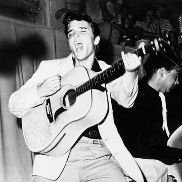 a man plays guitar and sings while standing, another man is in the background on the right