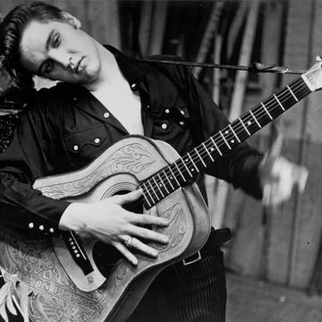 elvis presley leans against a wall as he holds an acoustic guitar