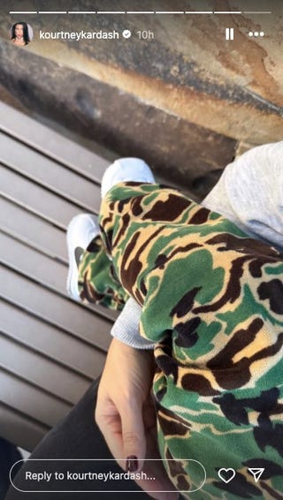 uma visão aproximada das pernas de uma pessoa vestindo calças camufladas e tênis brancos com uma mão descansando suavemente na perna