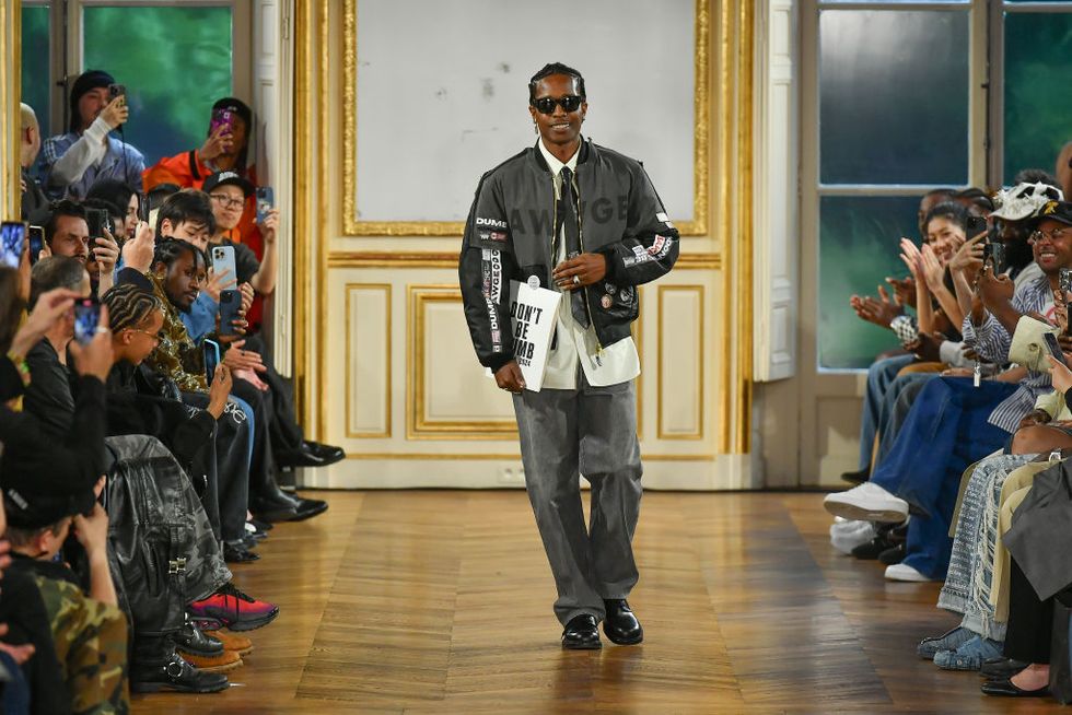 a$ap rocky walks down a runway as people watch from seats on both sides, he smiles and holds a book