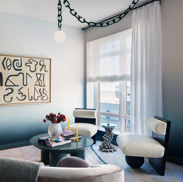 living room with ombre painted walls in shades from palest white blue to darker blue at the base along with a horizontal piece of art with black hieroglyphic type characters on it and a curved sofa in a off gray fabric and two armless chairs with column base and a matte blue round table at center