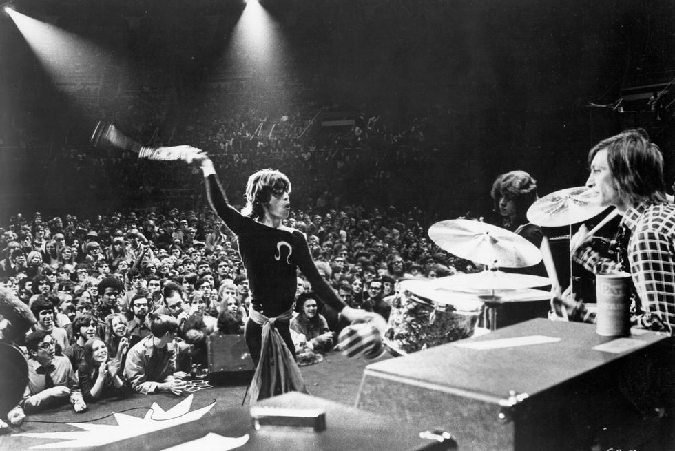 mick jagger whips something around his head while standing on a stage with drumsets and two other men, a crowd of people stands in the background
