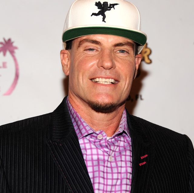 vanilla ice smiles at the camera, he wears a black pinstriped suit jacket with a purple checkered collared shirt and a white cap with a green bill