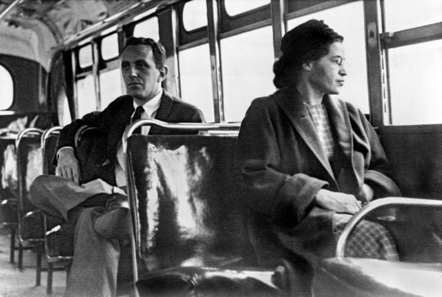 a black and white photograph of rosa park sitting in the seat of a bus, looking out the window, with a man sitting in the seat behind her