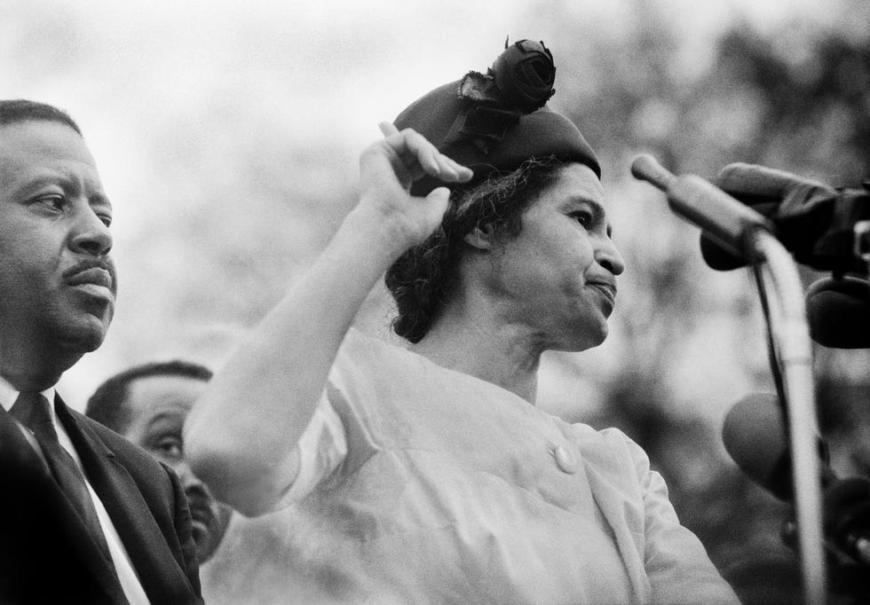 rosa parks stands behind microphones and points one finger in the air as she looks right