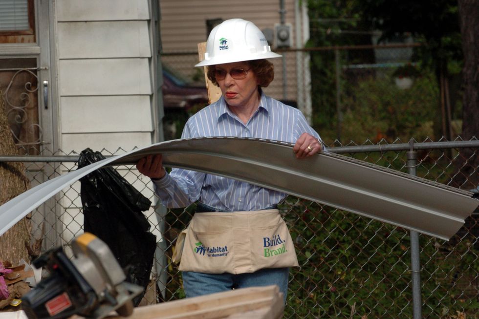 rosalynn carter holds a strip of house siding at a habitat for humanity work site, she is wearing a white hard hat, pink tinted sunglasses, a blue collard shirt with white strips, blue jeans and a fabric tool belt, in the left lower corner of the frame is a circular saw sitting on top of a work bench