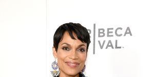 rosario dawson smiles at the camera with her hands on her hips while standing in front of a white backdrop, she wears a navy and white patterned dress with a navy bow tied at the waist