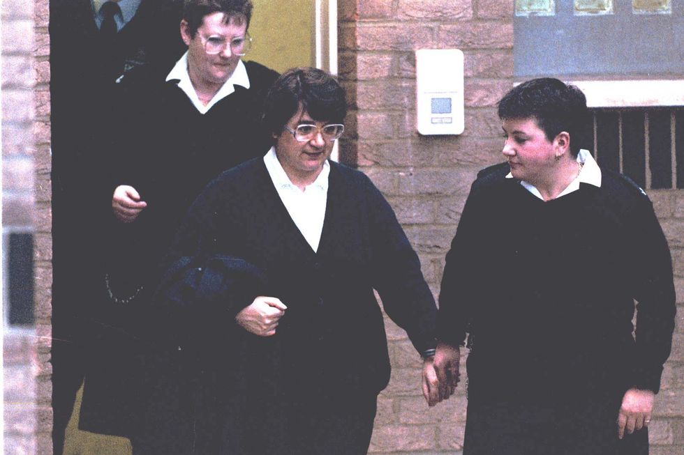 three women dress in black with white collared shirts walk outside a brick building, two women are handcuffed together