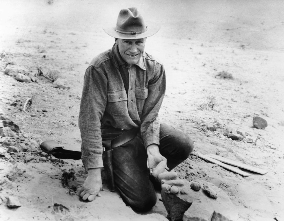 roy chapman andrews kneeling on the ground in the desert while holding dinosaur fossils and smiling for the camera