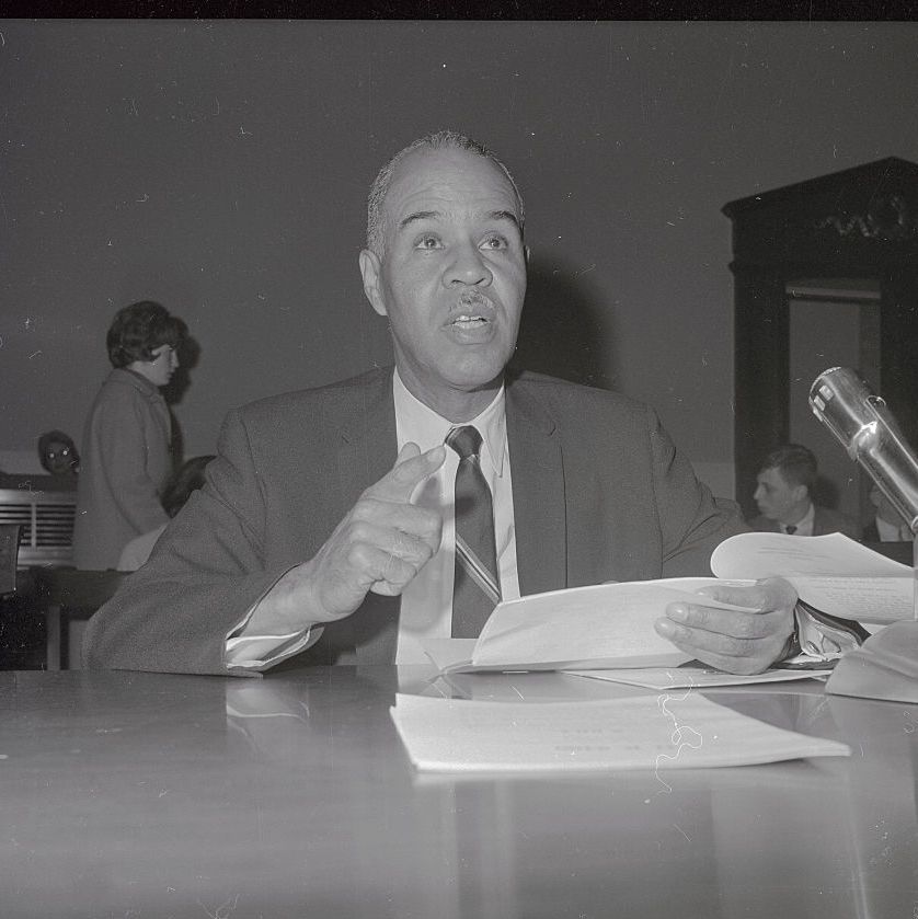 roy wilkins sits at a table and looks ahead while speaking and holding papers in one hand, he wears a suit jacket, tie and collared shirt, two people and a doorway are in the background
