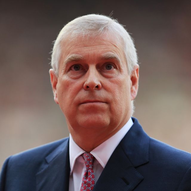 prince andrew looks past the camera with a neutral expression, he wears a blue suit jacket, white collared shirt and red patterned tie