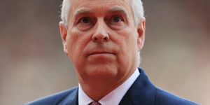 prince andrew looks past the camera with a neutral expression, he wears a blue suit jacket, white collared shirt and red patterned tie