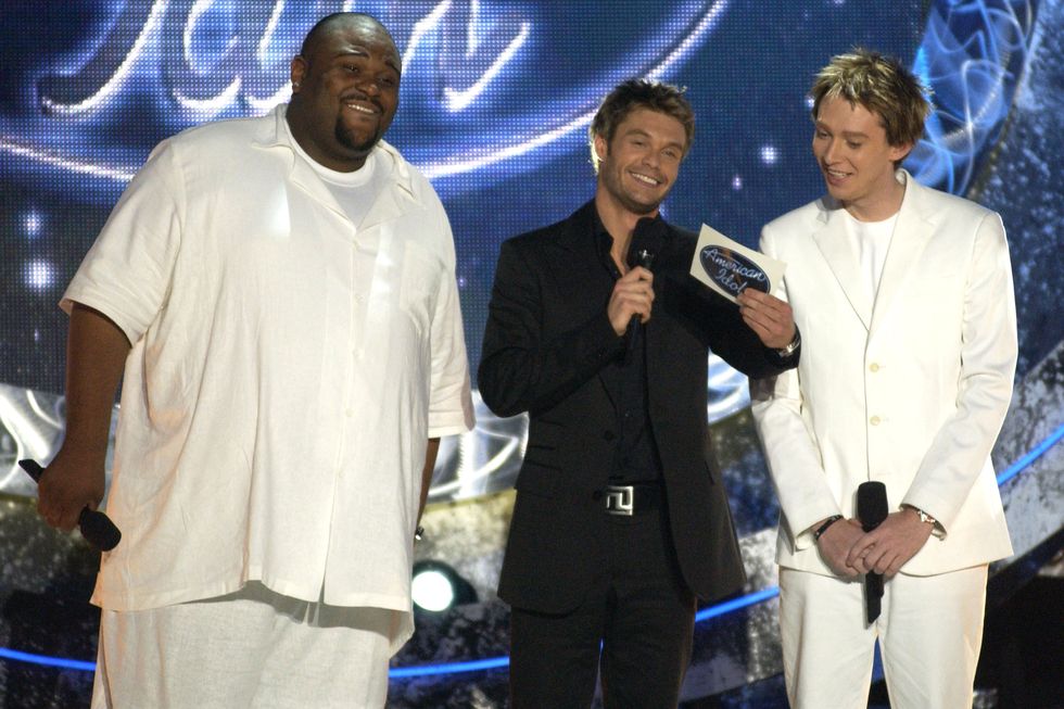 ryan seacrest looking at a cue card and holding a microphone between american idol contestants ruben studdard and clay aiken