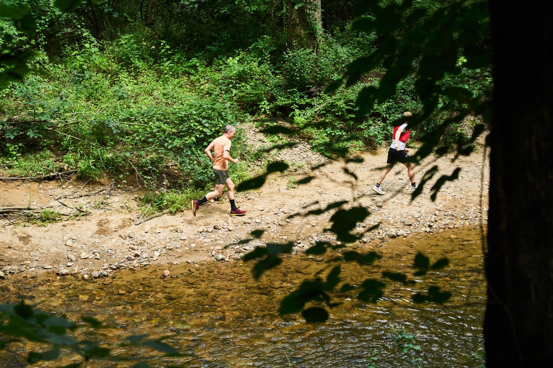 two runners on a dirt path next to a stream in a forested area