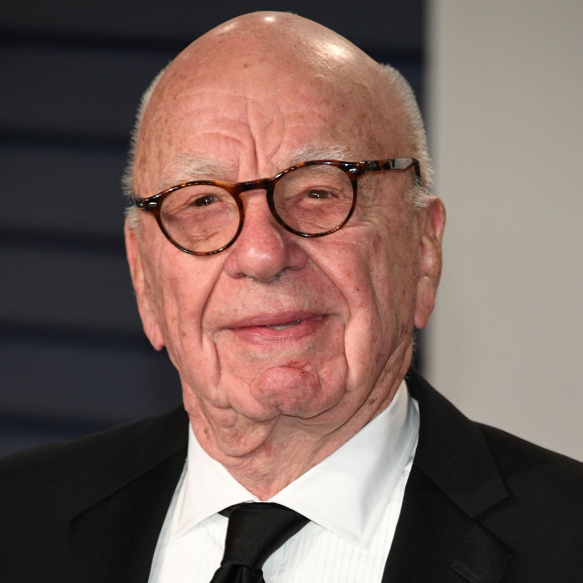 rupert murdoch looks at the camera with a slight smile, he is wearing a black suit jacket, white collard shirt, black tie, and brown tortoise shell glasses