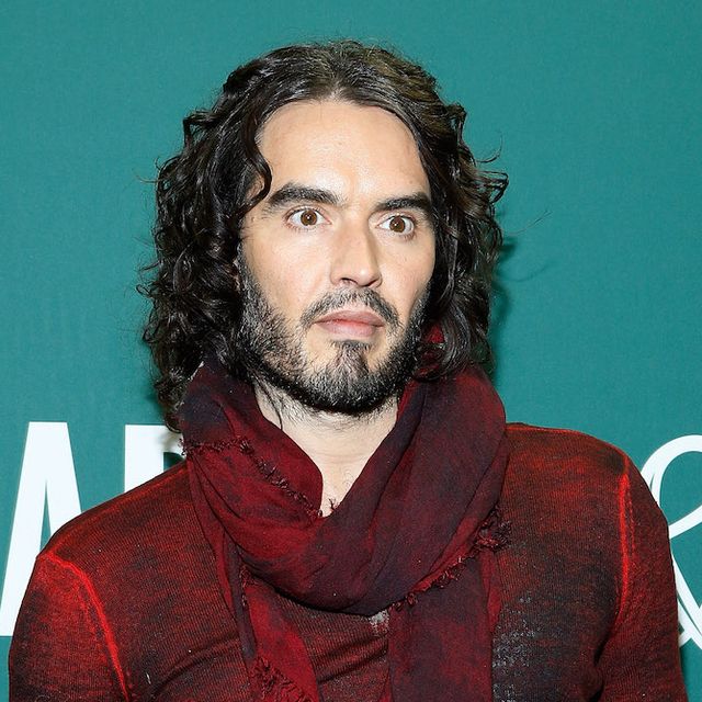 russell brand poses in a red top against a turquoise background at an event for his book launch
