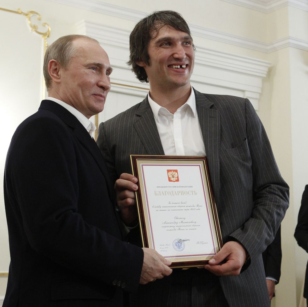 vladimir putin and alex ovechkin hold a framed certificate and pose for a photo together, both men wear suit jackets and white collared shirts and smile as they look right of the camera