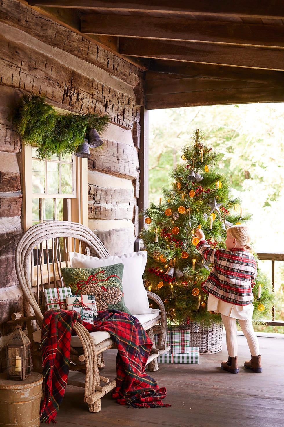 christmas tree on porch with small blonde girl in plaid jacket hanging an ornament