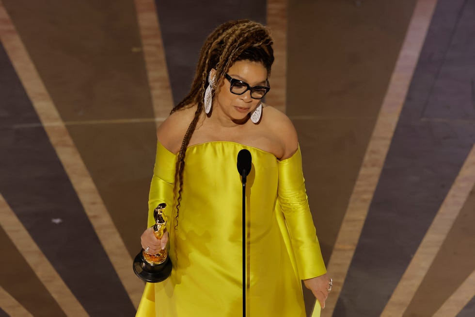ruth e carter, wearing a yellow dress, white earrings, and black glasses, holds an academy award and speaks into a microphone on a stage