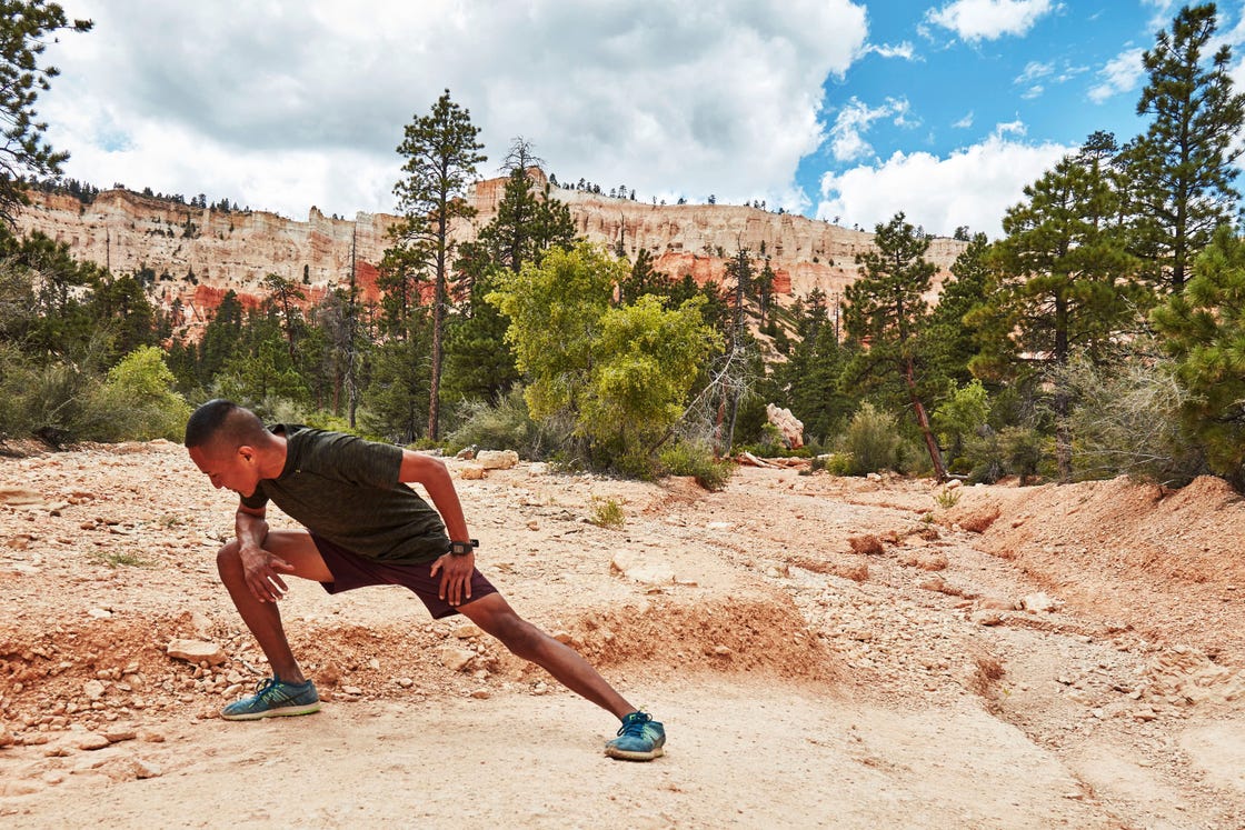 a man stretching on a dirt trail with trees and a mountain in the background