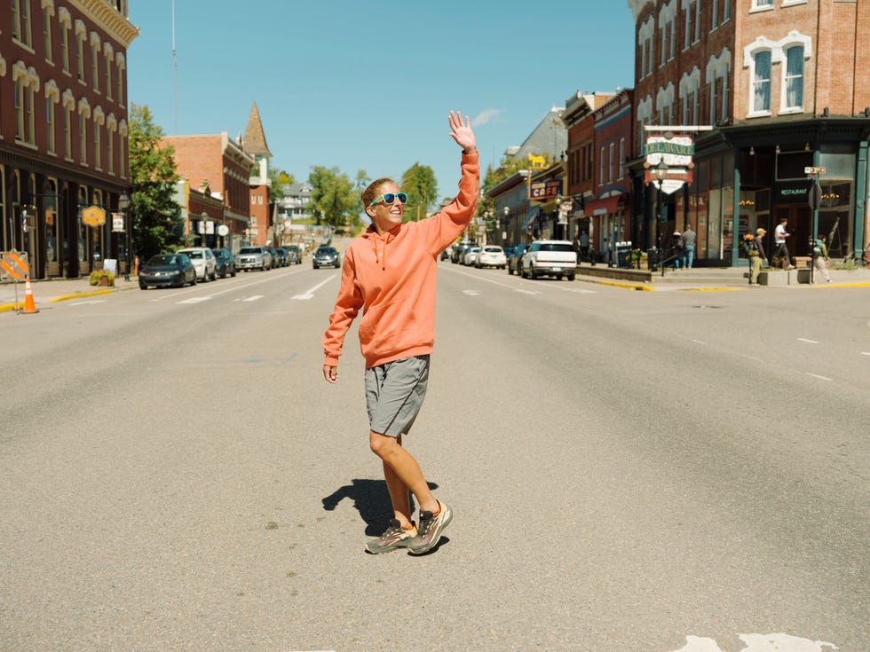 a woman wearing a hoodie waves while crossing a street
