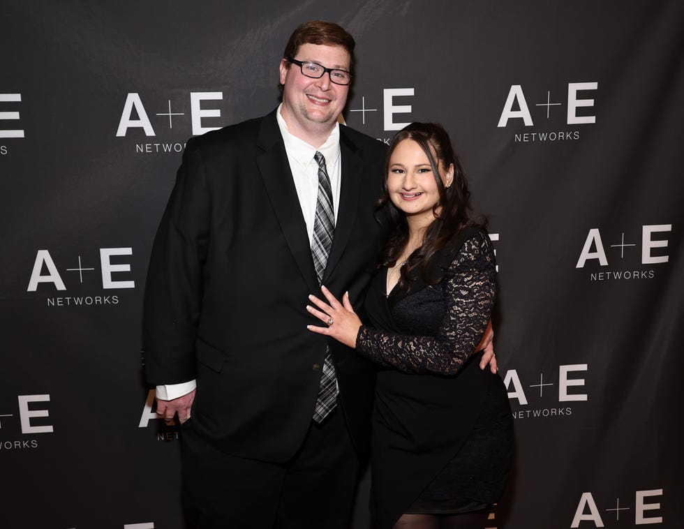 ryan anderson and gypsy rose blanchard stand in front of a black background with white written logos and smile at the camera, he wears a black suit with a white collared shirt and tie, she wears a black vneck dress with long sleeves and rests on hand on his chest
