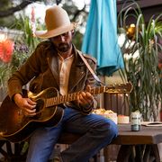 ryan bingham sitting on picnic table playing guitar with ranch rita canned cocktail on table