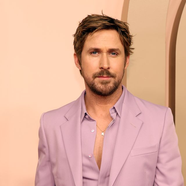 ryan gosling looks at the camera, he wears a light purple suit jacket with a matching collared shirt that is partially unbuttoned and a silver necklace