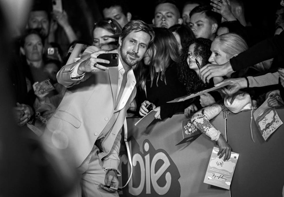 ryan gosling holds out of phone and takes a selfie with fans standing behind a fence with a barbie poster, he wears a suit