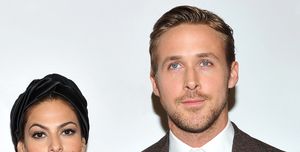 eva mendes and ryan gosling pose for a photo, she wears a sheer lace dress, black head wrap and dangling earrings, he wears a white collared shirt and a brown blazer