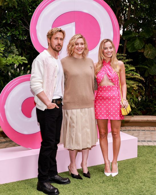 ryan gosling, greta gerwig, and margot robbie pose for a photo while standing in front of a large pink b in the barbie font, gosling wears a white shirt, light colored cardigan, and black pants, gerwig wears a camel colored sweater and tan pleated skirt, robbie wears a pink halter dress with white polka dots