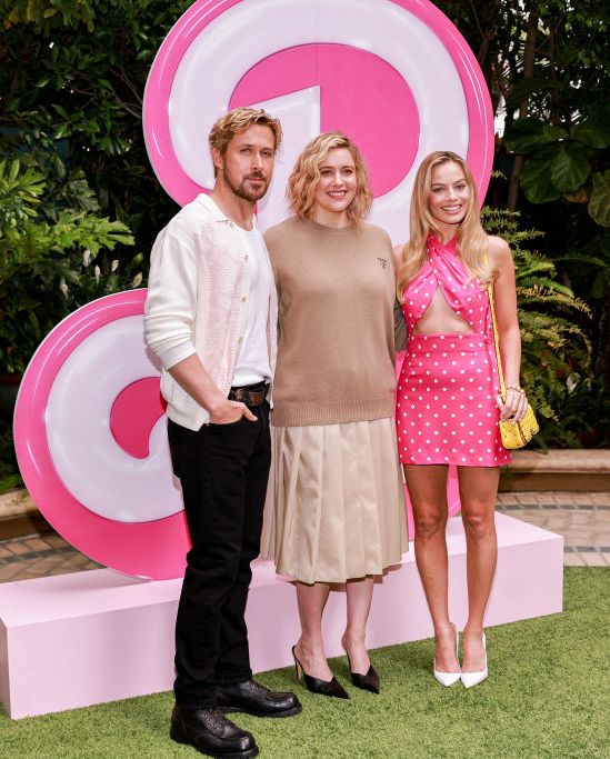 ryan gosling, greta gerwig, and margot robbie pose for a photo while standing in front of a large pink b in the barbie font, gosling wears a white shirt, light colored cardigan, and black pants, gerwig wears a camel colored sweater and tan pleated skirt, robbie wears a pink halter dress with white polka dots