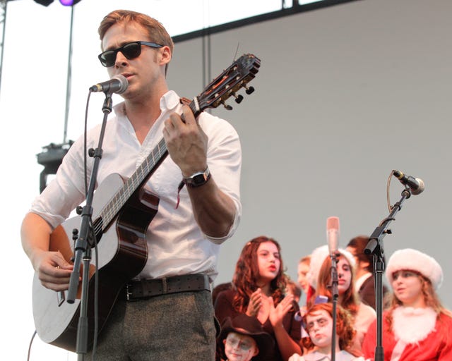 ryan gosling singing into a microphone and playing guitar on a stage, he wears sunglasses, a white collared shirt and gray pants with a belt
